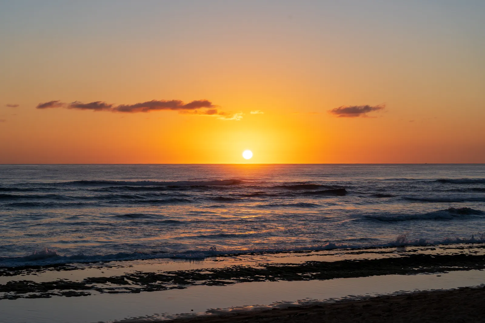 Disciplined minimal sunset: sun sitting on the horizon, a single cloud band, and a dark reef ledge anchoring the bottom third