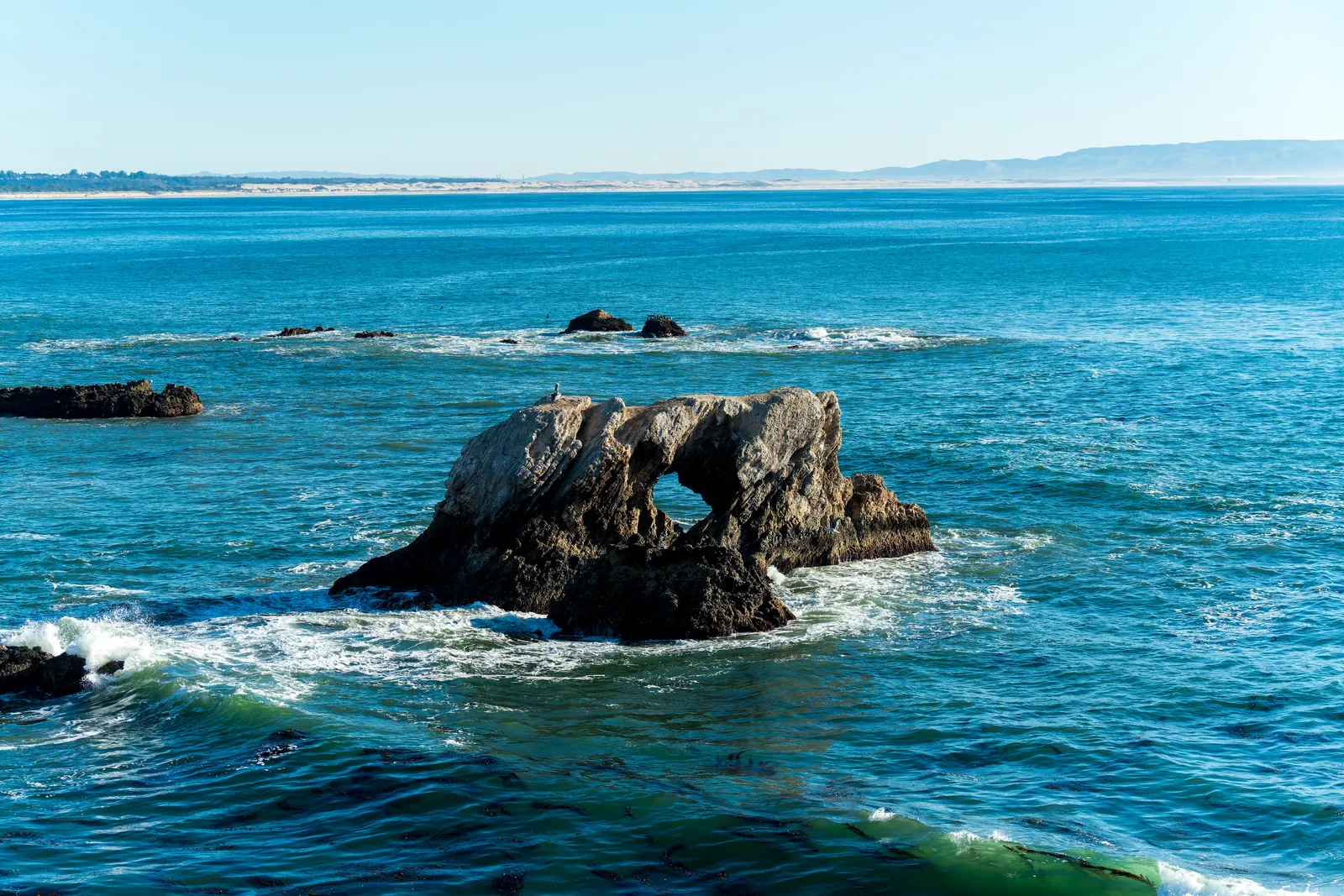 Sea arch isolated in turquoise water with surf wrapping its base and a distant headland setting the far plane. Clean subject separation, good water texture, no signage or structures. Competent coastline work that holds beside the published set