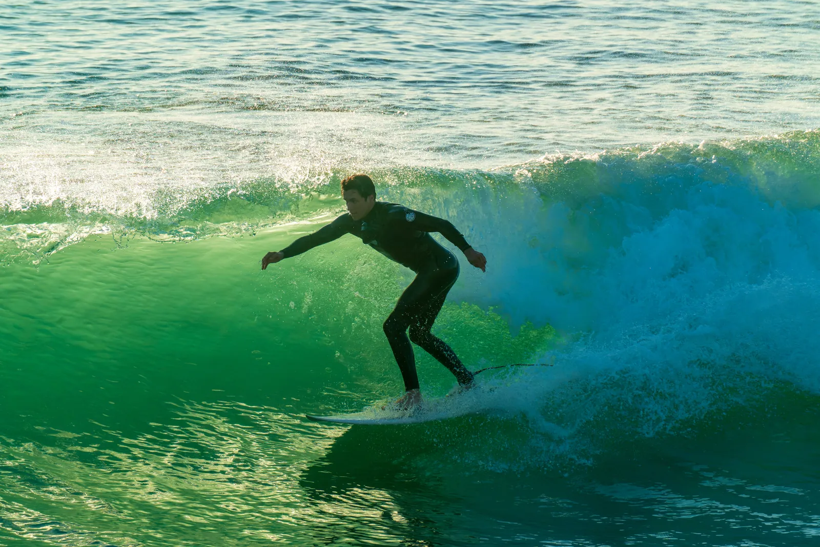 Surfer low and crouched on a clean green wave face, backlit water going translucent behind him, whitewater feathering along the lip. Real surf photography: single isolated subject, uncluttered water, strong colour separation between the figure and the wave. The one frame here that would hold beside published coastline work