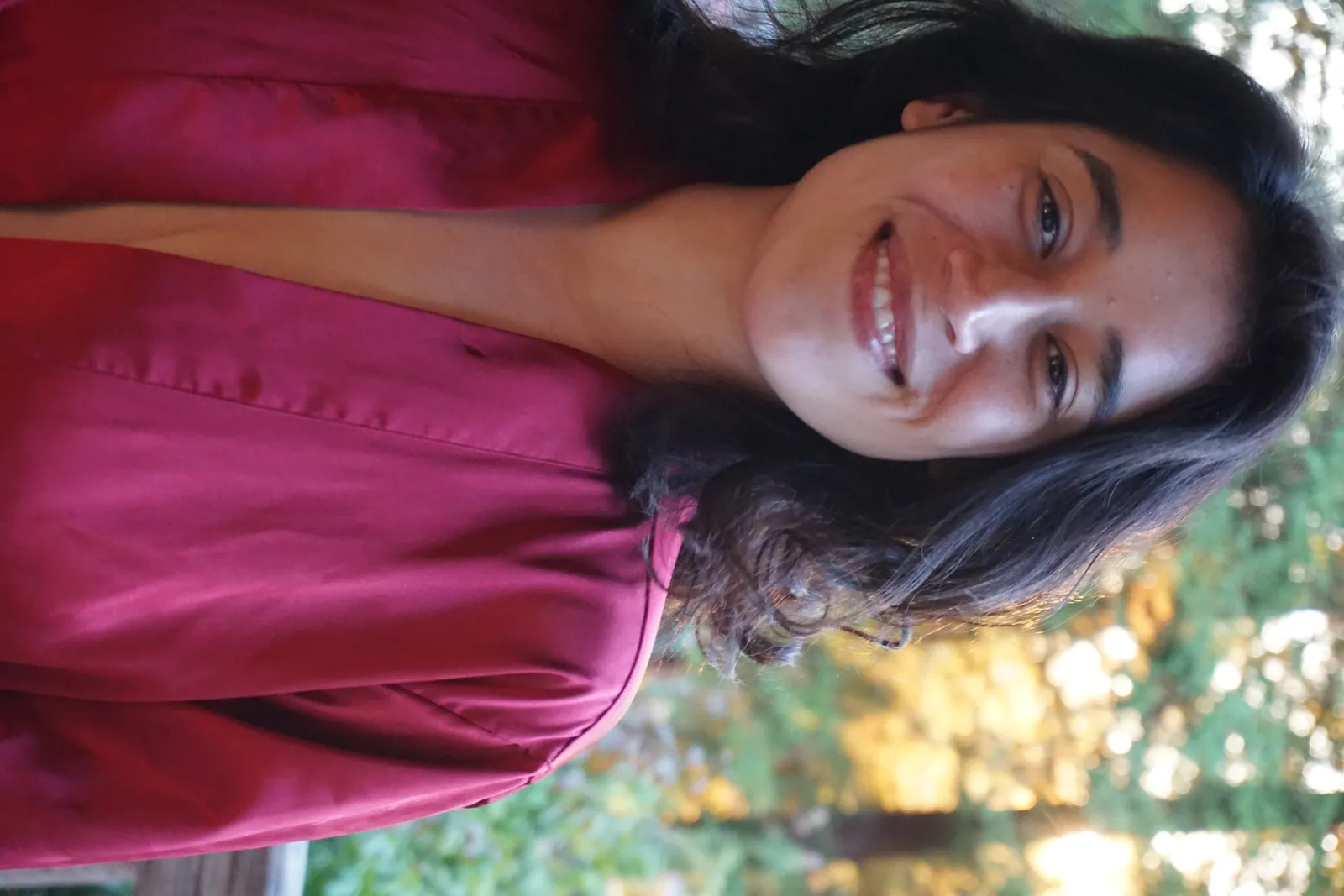 The best straight portrait in the batch: sharp on the eyes, warm open smile that is flattering rather than mid-laugh, golden late-light bokeh of redwoods behind, and the magenta robe separating cleanly from the background. Chosen over 0087 from the same sitting, which has a weaker expression and a cluttered deck behind her
