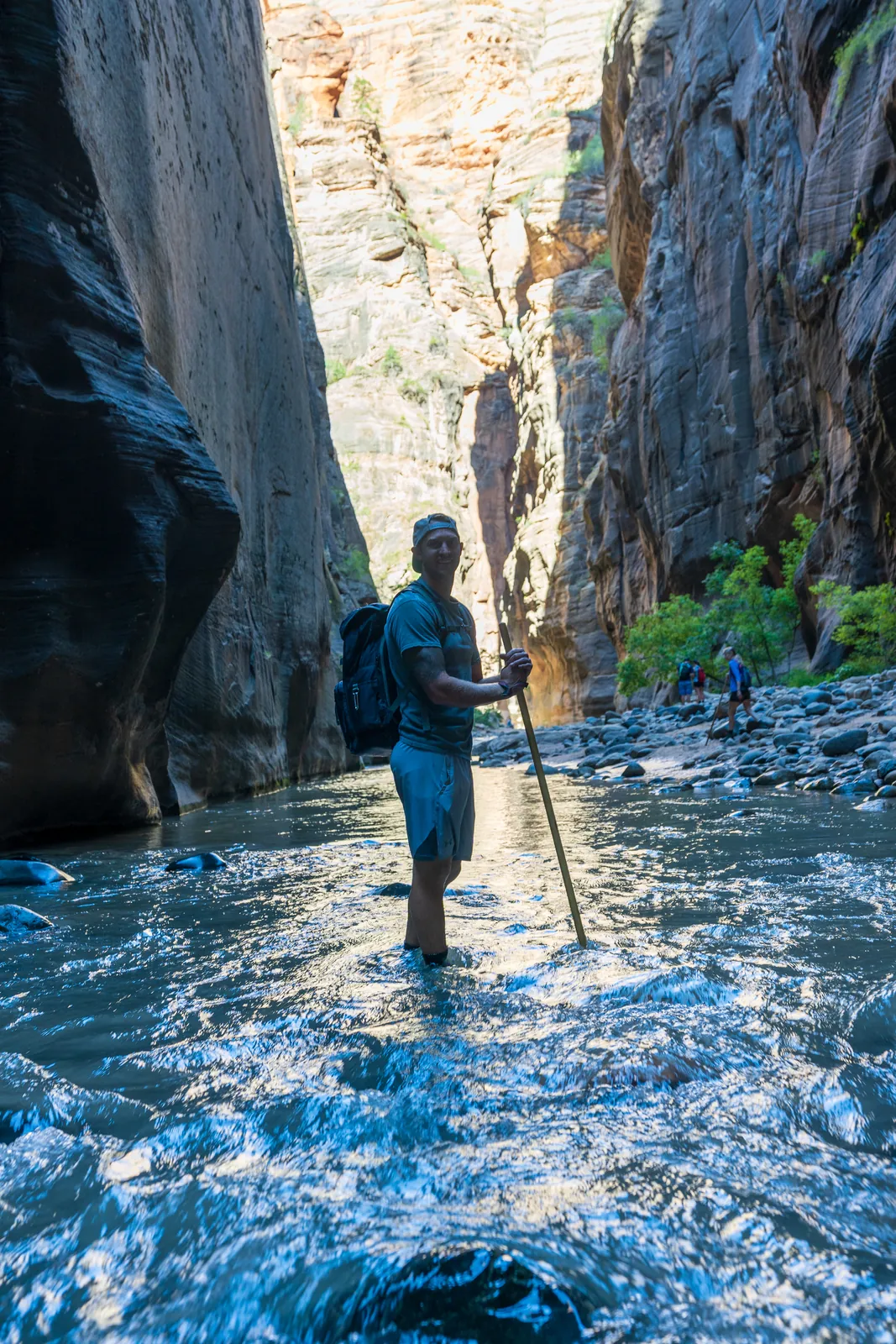 Lone hiker mid-river in the slot canyon, rim-lit against dark walls with sunlit water swirling in the foreground. The figure reads as form rather than a recognizable face, and the canyon dwarfs him